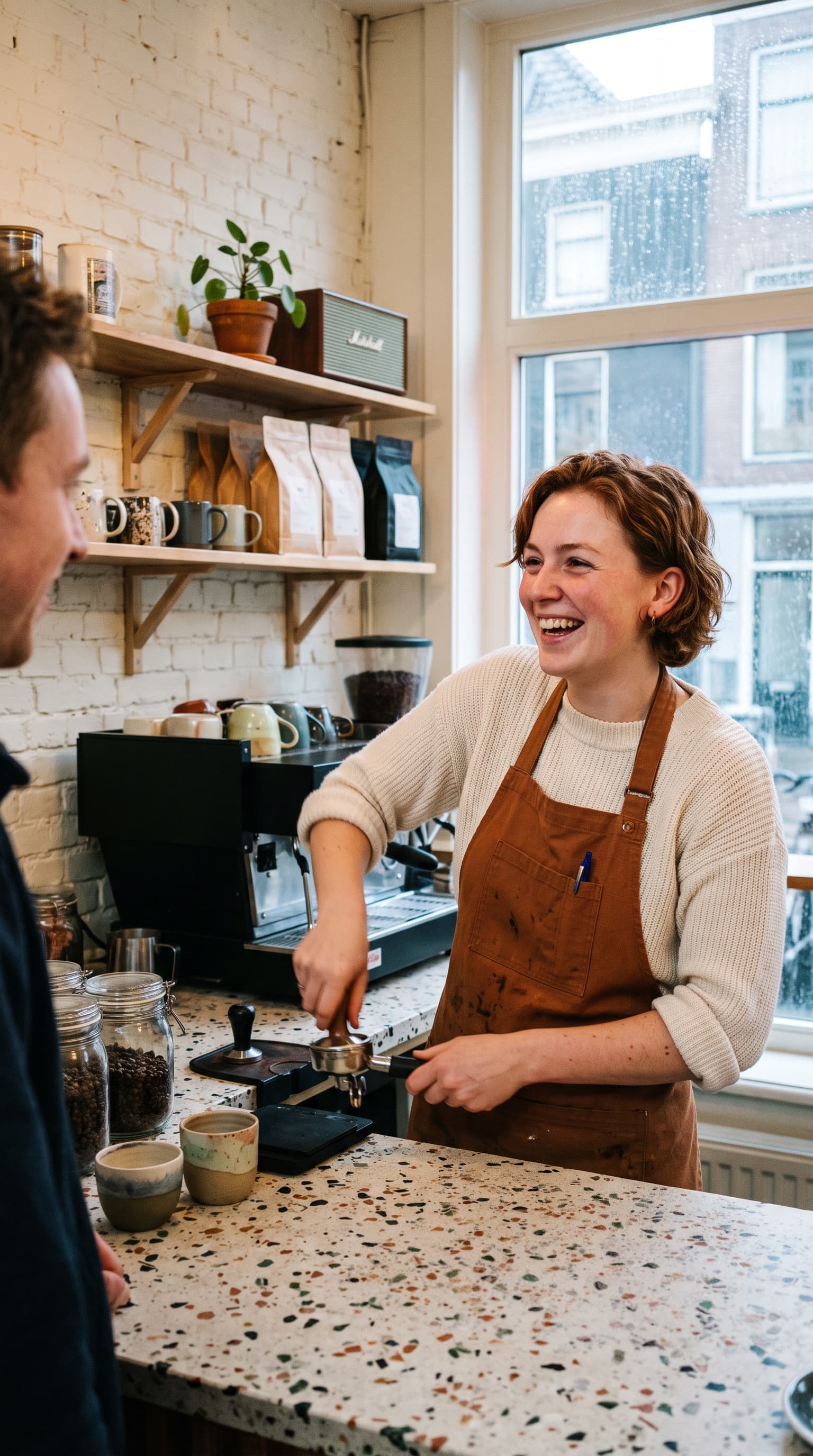 Een lachende barista met een bruin schort die espresso aandrukt achter een terrazzo toonbank, planken met kopjes en koffiezakken tegen een witte bakstenen muur, een hoog raam op de straat.